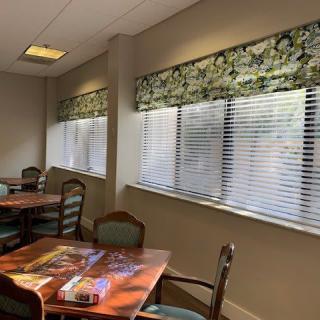 A cozy room with tables and chairs, featuring large windows with blinds.