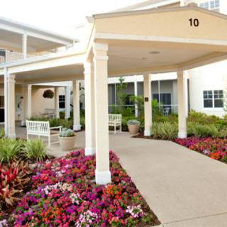A well-maintained building entrance with colorful flower beds lining a concrete pathway, featuring white columns and a covered entryway.