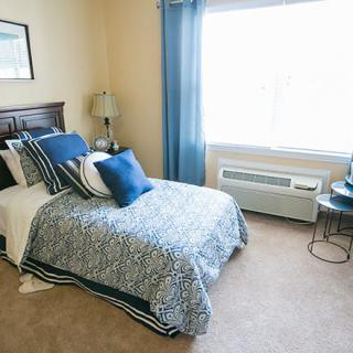 A cozy bedroom with a single bed adorned in blue decor, featuring a lamp and a large window.