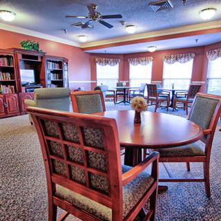 A cozy dining area with tables and chairs, featuring shelves and large windows with curtains.