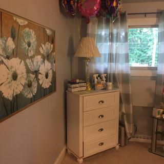 A cozy bedroom corner featuring a dresser with a lamp and balloons, alongside floral artwork and a window with curtains.