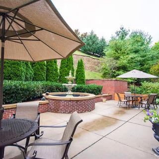 A tranquil outdoor patio area featuring tables and chairs under large umbrellas, with a decorative fountain surrounded by lush greenery.