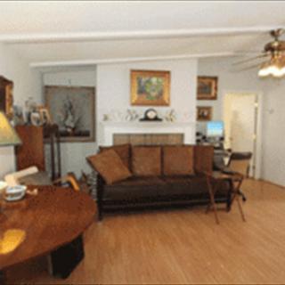 A cozy living room with a brown sofa, wooden coffee table, and various artworks adorning the walls. A ceiling fan hangs above, and a computer desk is situated in the corner.
