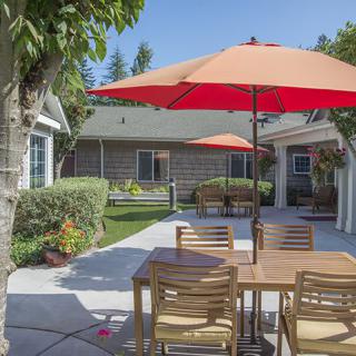 A sunny patio with wooden outdoor furniture and red umbrellas, surrounded by a well-maintained garden.