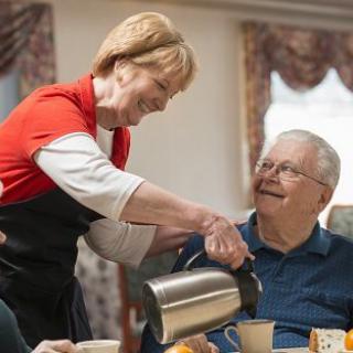 A caregiver pours coffee for a smiling elderly man in a cozy indoor setting.