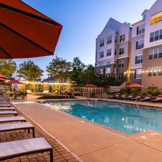 An outdoor swimming pool surrounded by sun loungers and umbrellas at an apartment complex during the evening.