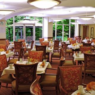 An elegant dining room with neatly arranged tables and chairs, surrounded by large windows offering a view of the greenery outside.