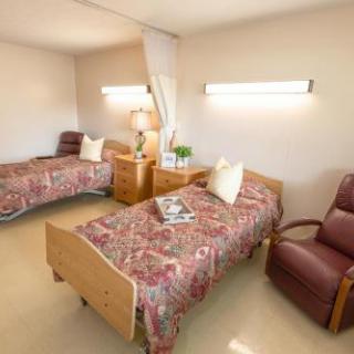 A shared hospital room with two beds, each adorned with floral bedspreads, accompanied by bedside tables and a comfortable chair.