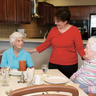 A caregiver engages warmly with two elderly women at a dining table in a senior living community.