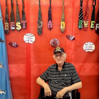 An elderly man sitting in a wheelchair with a display of ties behind him, celebrating 'Best Dad Ever'.