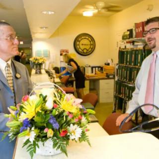 Two people in business attire having a conversation at a reception desk with a flower arrangement.