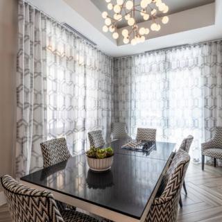 A modern dining room featuring a sleek black table, patterned chairs, and a striking chandelier, complemented by geometric curtains.