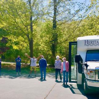 A group of people standing near a bus in a sunny, tree-lined area.