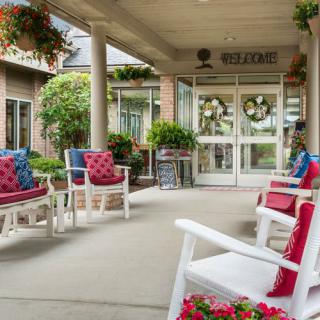 A welcoming porch area with rocking chairs adorned with decorative pillows, surrounded by plants at the entrance of a building.