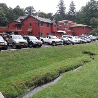 A view of red buildings with a parking lot filled with various cars, set against a lush green landscape and a small stream.