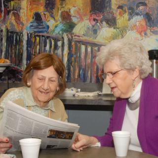 Two elderly women enjoying a conversation over a newspaper in a cafe setting.