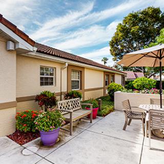 A cozy residential patio featuring outdoor seating with benches and a table under an umbrella, surrounded by potted plants and greenery.