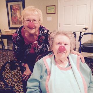 Two elderly women are smiling, with one playfully wearing a red clown nose, indoors.