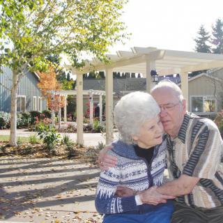 An elderly couple shares a tender moment in a garden setting.