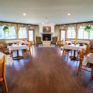 A cozy dining room with neatly arranged tables and chairs, featuring large windows and a central fireplace.