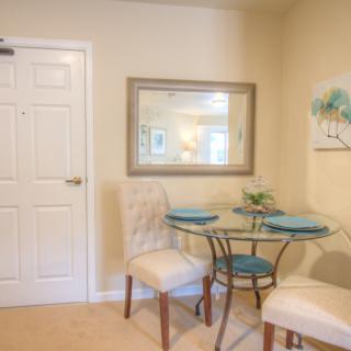 A cozy dining area featuring a glass round table with two upholstered chairs, complemented by a wall mirror and artwork.