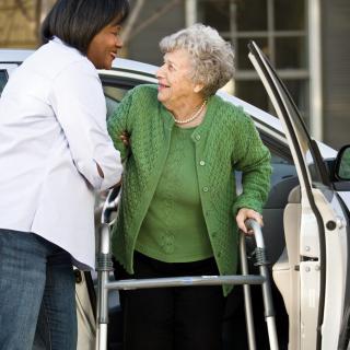 A caregiver assists an elderly woman with a walker as she gets out of a car.