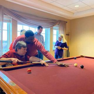 A group of people playing billiards indoors, with one person assisting another in making a shot.