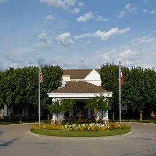 A building with flags in front, surrounded by trees and a landscaped roundabout under a partly cloudy sky.