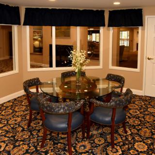 A cozy dining room with a round glass table and upholstered chairs, surrounded by large windows and decorated with a patterned carpet.