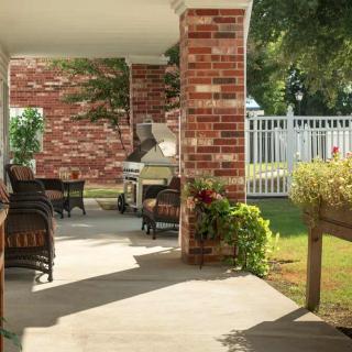 A cozy outdoor patio area with brick columns, comfortable seating, a grill, and lush greenery.