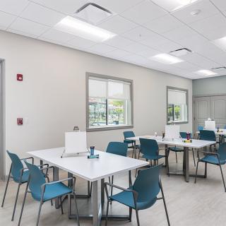 A modern office cafeteria with tables and chairs, featuring bright lighting and large windows.