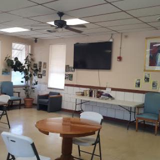 A cozy community room with a circular table, chairs, a television on the wall, and decorative plants.