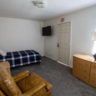 A minimalist bedroom with a single bed, armchair, dresser, and wall-mounted television.