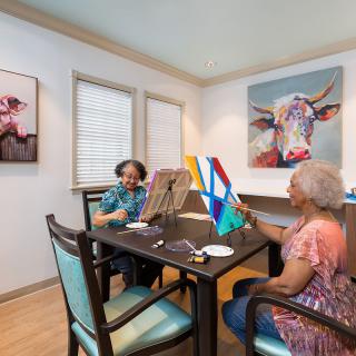 Two seniors enjoying a painting session indoors, surrounded by colorful artwork.