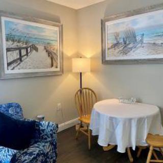 A cozy living room corner with two paintings, a small round table with a white tablecloth, wooden chairs, and a blue patterned armchair.