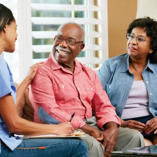 A caregiver converses with an elderly couple in a home setting, offering support and care.