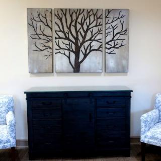 A triptych tree painting on a wall above a dark cabinet, flanked by two patterned chairs.