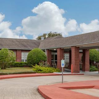 A brick building with a covered entrance and landscaped gardens under a blue, cloudy sky.