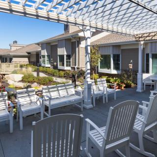 A sunny outdoor patio with chairs and benches under a pergola, surrounded by a garden.