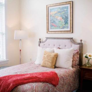 A cozy bedroom with a neatly made bed, framed painting above the headboard, and a floor lamp beside the bed.