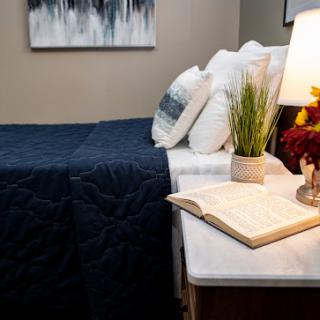 A cozy and inviting bedroom setup featuring a navy quilt, decorative pillows, a stylish lamp, and an open book on the nightstand.
