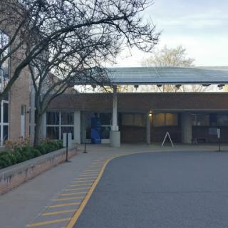 A view of the entrance to a building with a parking area, surrounded by trees.