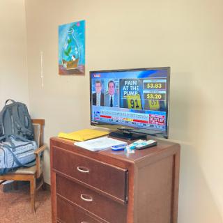 A cozy sitting area with a television displaying news, next to a wooden dresser and wall art.