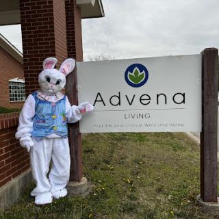 A cheerful Easter bunny character poses in front of the Advena sign.