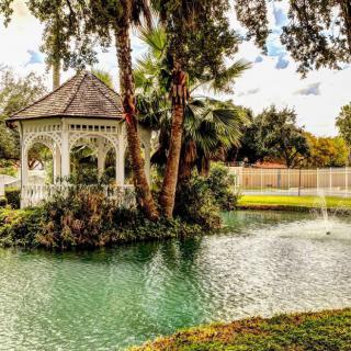 A tranquil garden scene featuring a gazebo by a pond with a fountain, surrounded by lush greenery.