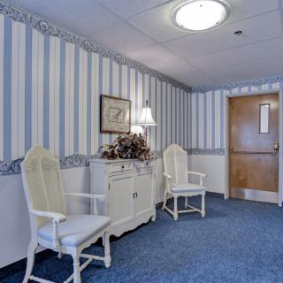 A cozy waiting area featuring two white chairs, a lamp on a dresser, and striped wallpaper.