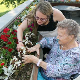 A younger woman assists an elderly woman with gardening, both smiling as they tend to the flowers together.