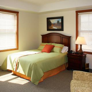 A cozy bedroom with a neatly made bed, wooden nightstand, and windows allowing natural light.