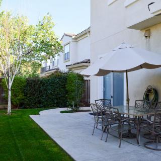 A well-maintained backyard with a patio featuring outdoor furniture and a large umbrella, surrounded by a lush green lawn and trees.