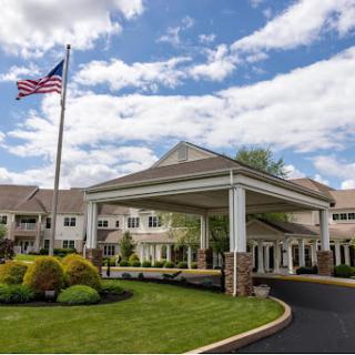 A welcoming entrance to a residential facility, adorned with a flag and surrounded by well-maintained landscaping under a cloudy sky.